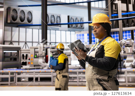 Female engineer in smart manufacturing plant monitors production line with tablet. The scene highlights industrial automation with robotic arms and solar panels, ensuring optimal factory performance. 127613634