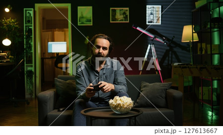 Gamer on couch in apartment engaging in online competitive multiplayer tourney using gamepad. Man in home theatre playing engaging videogame with controller, eating popcorn, camera B 127613667