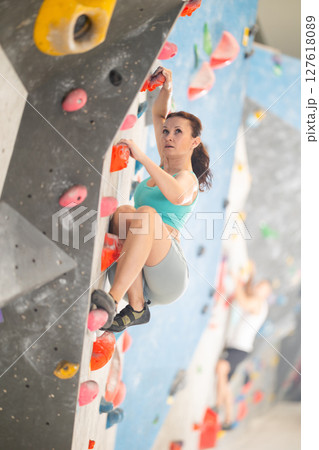 Woman in comfortable athletic clothing moving carefully on bouldering wall clinging to colorful hand and foot holds 127618089
