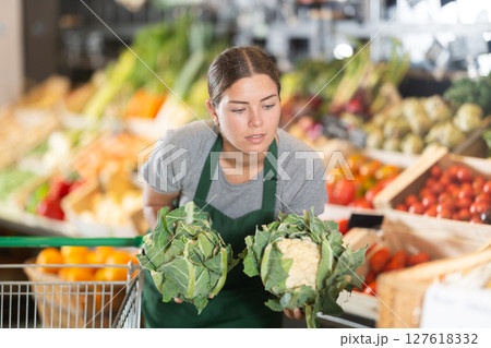 Girl supermarket worker with cauliflower in hands examines product Girl supermarket worker with cauliflower in hands examines product 127618332