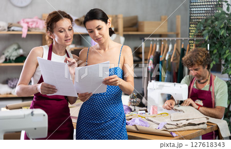 Two female dressmakers discussing dress sketches on paper. Man works on sewing machine. Two female dressmakers discussing dress sketches on paper. Man works on sewing machine. 127618343