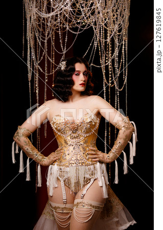 Cabaret dancer in vintage corset costume standing under chandelier on dark stage Cabaret dancer in vintage corset costume standing under chandelier on dark stage 127619845