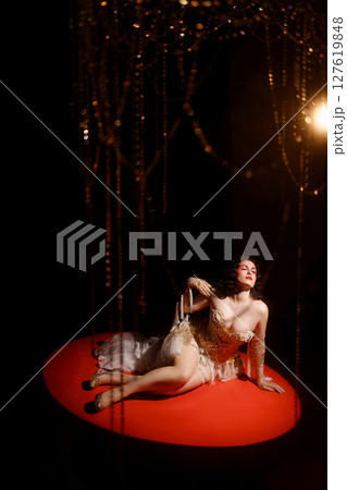 Woman in vintage corset lying on red stage floor under hanging beads on dark background 127619848
