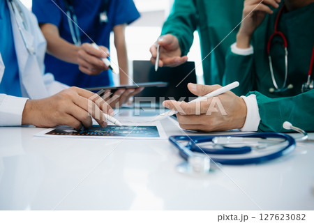 Medical team having a meeting with doctors in white lab coats and surgical scrubs seated at a table discussing a patients working online using computers 127623082