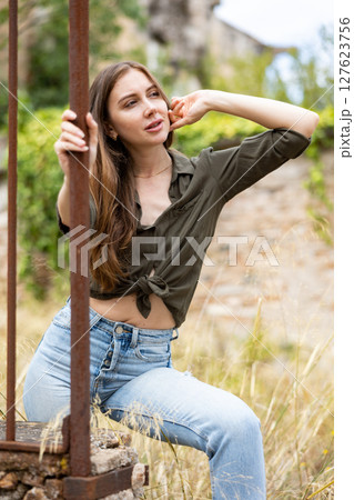 Woman in jeans and shirt posing on camera on street. Female model with blond hair looking to camera and standing near wrought-iron fence Woman in jeans and shirt posing on camera on street. Female model with blond hair looking to camera and standing near wrought-iron fence 127623756