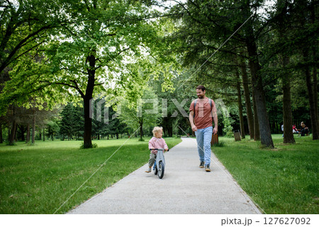 Toddler riding balance bike with dad cheering behind Toddler riding balance bike with dad cheering behind 127627092