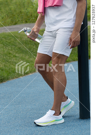 Legs of sporty man wearing white clothes holding water bottle during outdoor exercise break at urban sport ground after workout. Outdoor air training. 127628937