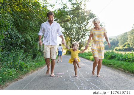 Parents helping litthe girl with hopscotch during warm summer day. Parents helping litthe girl with hopscotch during warm summer day. 127629712