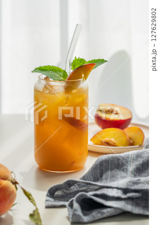 Peach drink or tea with ice, mint in a glass on a white table near window with fresh fruits 127629802