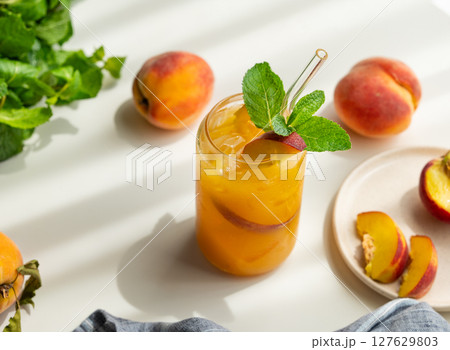 Peach drink or tea with ice, mint in a glass on a white table with fresh fruits and sunlight. 127629803