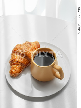 Coffee espresso in yellow cup and fresh croissant on a plate on white table near window Coffee espresso in yellow cup and fresh croissant on a plate on white table near window 127629810