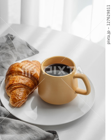 Coffee espresso in yellow cup and fresh croissant on a plate on white table near window 127629811