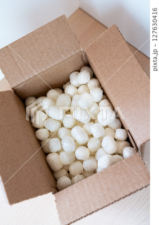 Open cardboard box filled with eco-friendly corn-based packing peanuts on a light wooden surface. Sustainable packaging for safe and conscious shipping, promoting zero-waste 127630416