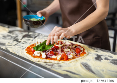 Cooker in apron working in the pizzeria kitchen and putting topping on the pizza Cooker in apron working in the pizzeria kitchen and putting topping on the pizza 127631160