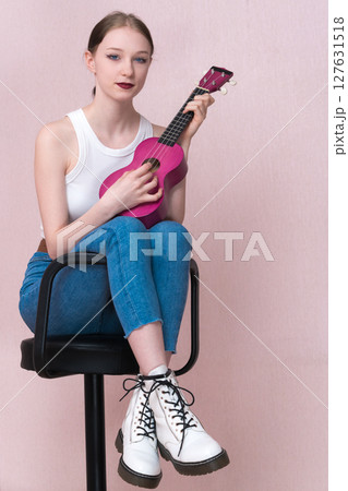 Young female musician strumming ukulele, sitting on bar stool and looking at camera while practicing musical instrument. Performer woman wearing white tank top, blue skinny jeans and white boots Young female musician strumming ukulele, sitting on bar stool and looking at camera while practicing musical instrument. Performer woman wearing white tank top, blue skinny jeans and white boots 127631518