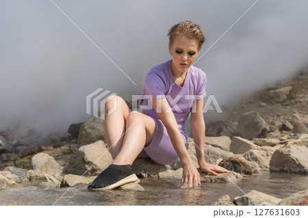 Woman tourist sits on rocks on bank of thermal stream in volcano crater and touches temperature of water with hand. Traveler against backdrop of clouds of steam and gas from hot springs and geysers 127631603