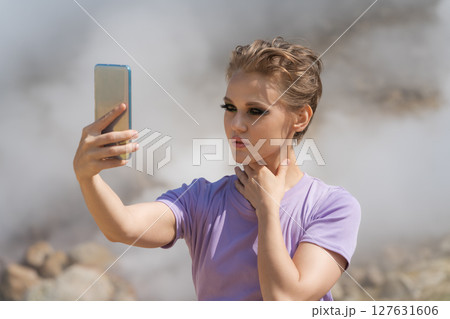 Beautiful female traveler using her cell phone to record self portrait to video call with friends in remote location against backdrop of steaming geysers in crater of an active volcano while traveling Beautiful female traveler using her cell phone to record self portrait to video call with friends in remote location against backdrop of steaming geysers in crater of an active volcano while traveling 127631606