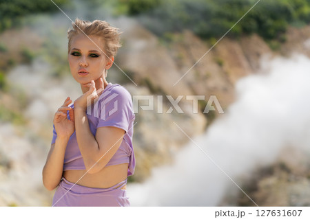 Fashion model looking at camera, standing against backdrop of volcano crater, with clouds of gas, pair of steaming geysers in background. Nature's beauty, plenty of copy space to add creative ideas 127631607