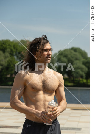 Shirtless fit brunette topless man with curly long hair holding a water bottle while standing outdoors on a sunny day against clear blue sky and lush green trees as nature background. Heat summer. 127632406