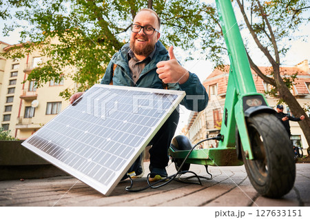 Happy man holds solar panel, for charging electric scooter in urban settings, giving thumbs up. Integration of solar power as sustainable energy source, promoting eco-friendly urban transportation. 127633151