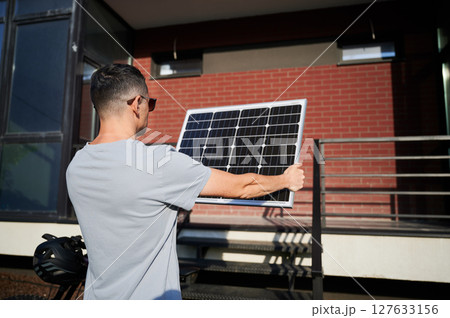 Back view of man holding solar panel toward the sun for charging electric mountain bike. Scene set against brick building. Back view of man holding solar panel toward the sun for charging electric mountain bike. Scene set against brick building. 127633156