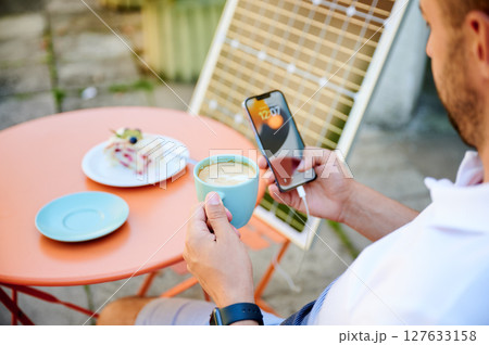 Young male drinking coffee and checking his phone while it charging. Focus on cup of coffee. Man using portable solar battery to charge his devices. Concept of using alternative energy everyday. Young male drinking coffee and checking his phone while it charging. Focus on cup of coffee. Man using portable solar battery to charge his devices. Concept of using alternative energy everyday. 127633158