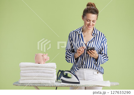 Smiling woman using phone near piggy bank and ironed towels 127633290