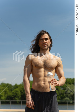 Brunette topless man with curly long hair holding water bottle looking at camera against clear blue sky and nature background. Heat summer. Brunette topless man with curly long hair holding water bottle looking at camera against clear blue sky and nature background. Heat summer. 127633716