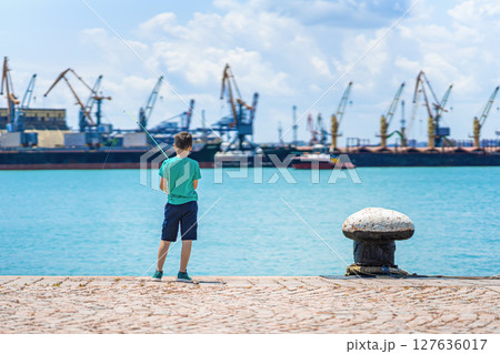 Boy fishing on sunny pier with industrial port in background, standing near turquoise water. Concept of fishing, childhood moments, peaceful outdoor leisure 127636017