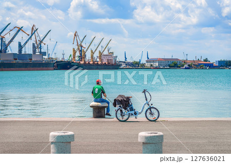 Man with fishing rod sitting on bollard near bicycle at harbor promenade, gazing over turquoise water toward industrial cranes. Concept of fishing, urban solitude, reflective outdoor moment Man with fishing rod sitting on bollard near bicycle at harbor promenade, gazing over turquoise water toward industrial cranes. Concept of fishing, urban solitude, reflective outdoor moment 127636021