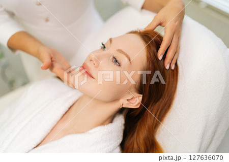 Close-up of beautician performing relaxing facial massage for young woman with long red hair, lying on massage table in white bathrobe, enjoying skincare treatment in modern beauty salon. 127636070