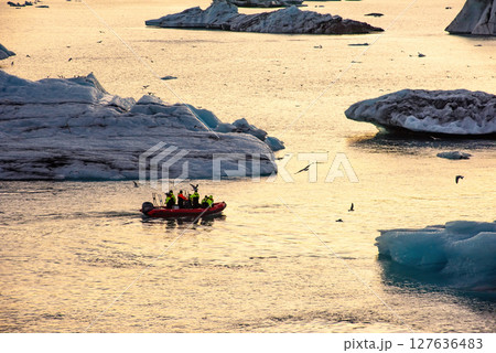 Group wearing bright jackets explores icebergs on an inflatable boat in a serene water setting during sunset, capturing a moment of adventure and exploration amidst icy landscapes Group wearing bright jackets explores icebergs on an inflatable boat in a serene water setting during sunset, capturing a moment of adventure and exploration amidst icy landscapes 127636483