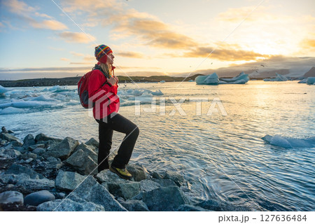 Woman in red jacket joyfully poses on rocky shore of a glacier lake in Iceland. Smiling broadly, she spreads her arms wide, capturing the beauty of the glowing sunset Woman in red jacket joyfully poses on rocky shore of a glacier lake in Iceland. Smiling broadly, she spreads her arms wide, capturing the beauty of the glowing sunset 127636484
