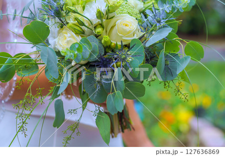 Bridal bouquet filled with white roses, greenery, purple accents held by bride in garden. 127636869