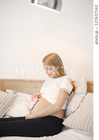Portrait of pregnant woman holding a little baby rose socks. Pregnant woman lie on a bed in living room. Blonde woman wearing white t-shirt and black trousers. Portrait of pregnant woman holding a little baby rose socks. Pregnant woman lie on a bed in living room. Blonde woman wearing white t-shirt and black trousers. 127637241