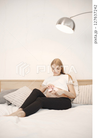 Portrait of pregnant woman holding a little baby rose socks. Pregnant woman lie on a bed in living room. Blonde woman wearing white t-shirt and black trousers. 127637242