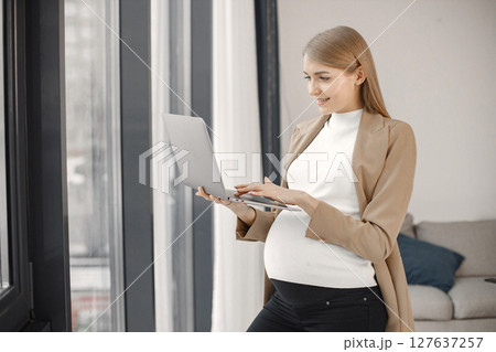 Portrait of pregnant woman typing on a laptop while standing near windows in modern office. Pregnant businesswoman using a laptop in her office. Blonde woman wearing beige jacket. 127637257