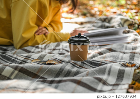 Takeaway coffee cup sits on plaid blanket in an autumn setting, with a person in yellow sweatshirt reading in the background Takeaway coffee cup sits on plaid blanket in an autumn setting, with a person in yellow sweatshirt reading in the background 127637934