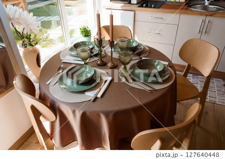 Cozy Dining Table Setting with Green Dishware, Candles, and Wooden Chairs in a Bright Minimalist Kitchen Interior Design, Earth Tones 127640448