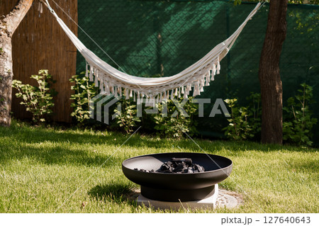 Cozy Backyard Retreat with White Hammock and Fire Pit Scene: Relaxing Outdoor Living, Green Garden, Summer Comfort, and Dark Metal Fire Bowl. 127640643