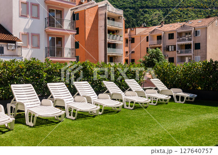 White lounge chairs on artificial grass rooftop. Urban sunbathing, summer relaxation, and outdoor leisure in residential neighborhood. 127640757