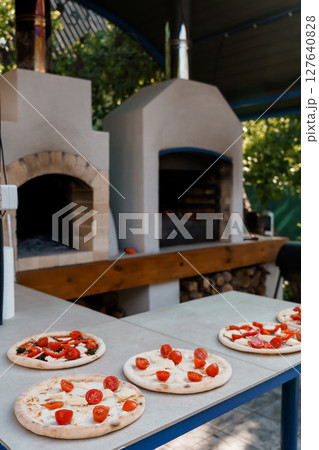 Rustic Outdoor Wood-Fired Pizza Oven and Prepared Pizzas: Warm Italian Cuisine, Stone Texture, Garden Setting, Summertime Food Photography 127640828