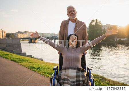 Senior woman in wheelchair walking with caregiver old man on road. Family couple man husband supporting taking care of paralyzed woman wife in chair for people with disability outdoor. Rehabilitation Senior woman in wheelchair walking with caregiver old man on road. Family couple man husband supporting taking care of paralyzed woman wife in chair for people with disability outdoor. Rehabilitation 127642515