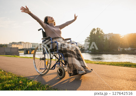 Happy senior woman in wheelchair raised hands enjoying freedom walking on road in city park outdoor. Old happy woman in chair for people with disability has a hope. Disability disease rehabilitation 127642516
