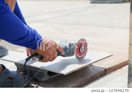 Construction site scene with a worker in blue clothing using an angle grinder to cut a gray tile, highlighting detailed craftsmanship and precision in tile preparation. Construction site scene with a worker in blue clothing using an angle grinder to cut a gray tile, highlighting detailed craftsmanship and precision in tile preparation. 127646270