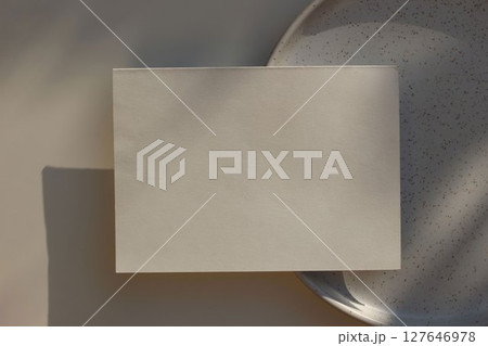 Summer minimal wedding stationery. Blank horizontal textured paper greeting card mockup, invitation. Speckled ceramic plate in sunlight, long shadows. Table background, shadows overlay. Flatlay, top 127646978