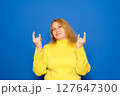 Delighted young ethnic female millennial with long dark hair in warm sweater and sunglasses, smiling brightly and showing rock gesture while listening to favorite song against yellow background 127647300