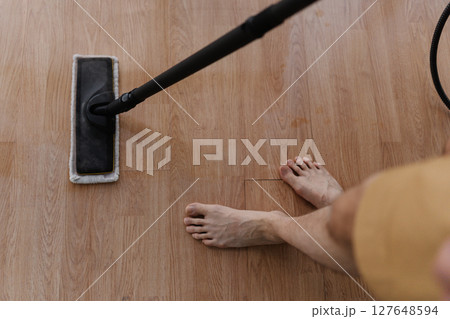 Top view of barefoot male cleaning hardwood floor with steam mop during house cleaning, close-up. Maintaining hygiene and cleanliness in home environment. Concept of cleaning and household. Top view of barefoot male cleaning hardwood floor with steam mop during house cleaning, close-up. Maintaining hygiene and cleanliness in home environment. Concept of cleaning and household. 127648594