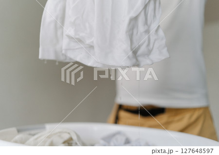 Cropped shot of unrecognizable man squeezing dripping wet white t-shirt over laundry basket full of soaking clothes, preparing laundry for washing machine, standing in living room at home. Cropped shot of unrecognizable man squeezing dripping wet white t-shirt over laundry basket full of soaking clothes, preparing laundry for washing machine, standing in living room at home. 127648597