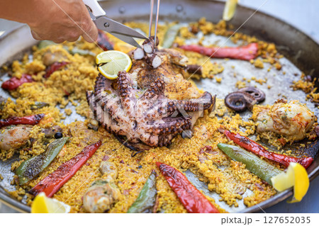 Seafood paella with whole octopus, peppers, lemon slices being served from metal pan at food festival. Concept of seafood, exotic dish, gourmet cooking on display Seafood paella with whole octopus, peppers, lemon slices being served from metal pan at food festival. Concept of seafood, exotic dish, gourmet cooking on display 127652035